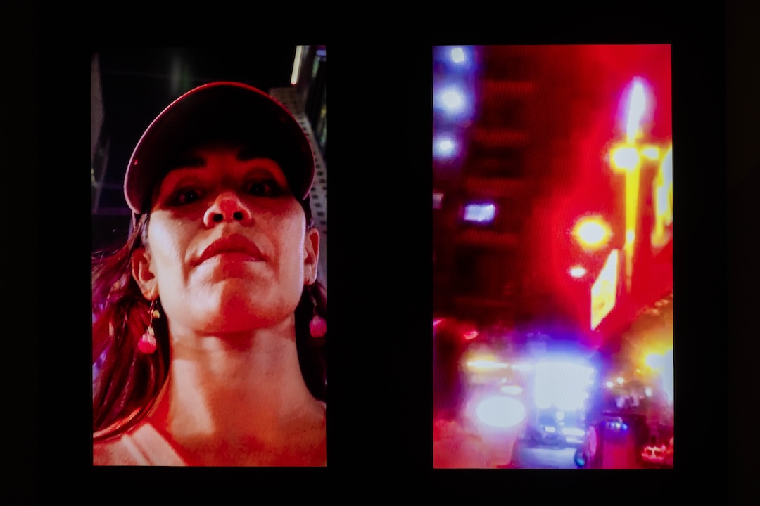 An image of two screens in a dark space. One screen shows the artist as she walks outside in her home city. One shows the cityscape at night.